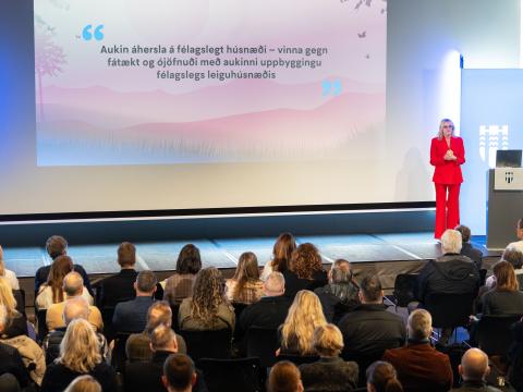 Heiða Björg Hilmisdóttir borgarstjóri í rauðri dragt. Stendur lengst til hægri á sviði, fjölmenni fylgist með henni tala í Tjarnarsal. Húsnæðisfundur. Texti á tjaldi á bakvið hana.