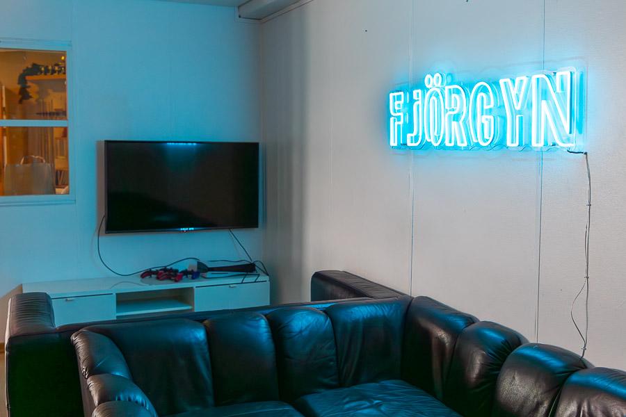 Black couches in front of the screen. On the wall is a lighted sign with the word Fjörgyn.