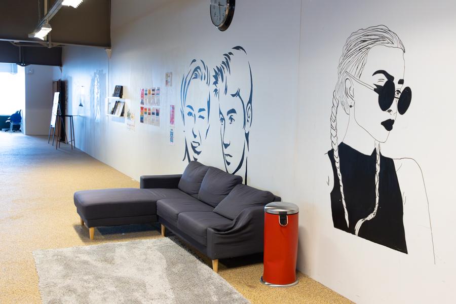 Gray sofa, light area rug, and red garbage can. Pictures of celebrities in black-and-white are painted on the wall behind.