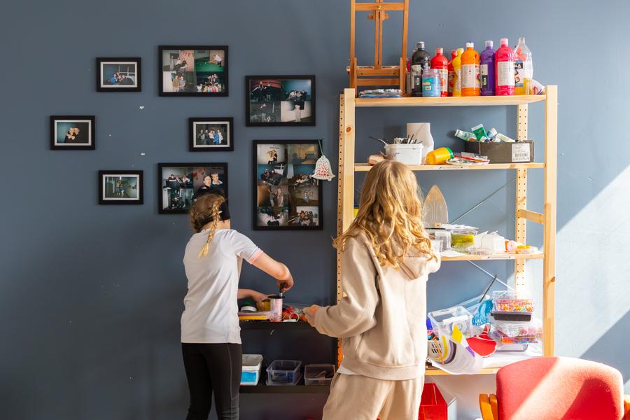Children stand in front of a shelf with paint and various craft supplies. In the background, a gray wall is decorated with pictures. 