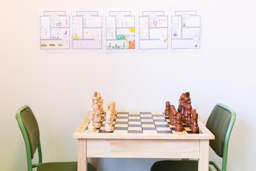 A chess table and two green chairs. Pictures on a wall.