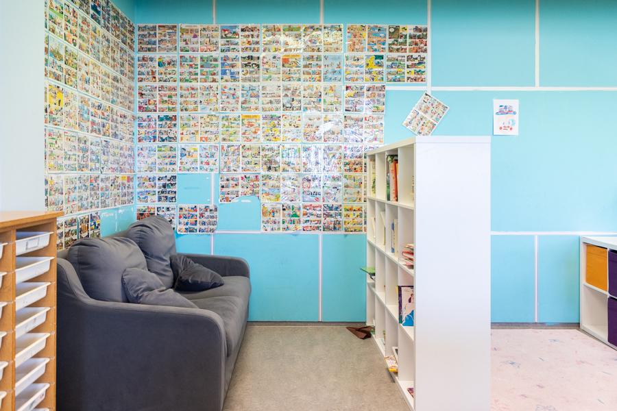 A corner with a gray couch and a bookshelf. Countless comic book pages are pasted on a wall. 