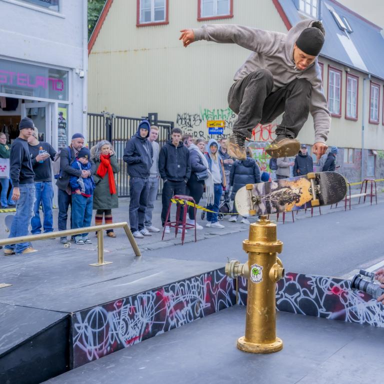 A skateboarder shows off his skills on Hverfisgata, jumping over a golden fire hydrant, with people watching in the background