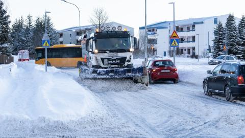snjómoksturstæki að moka á götu. Snjór, hús og bílar í kring.