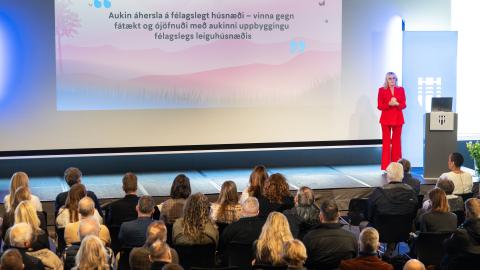 Heiða Björg Hilmisdóttir borgarstjóri í rauðri dragt. Stendur lengst til hægri á sviði, fjölmenni fylgist með henni tala í Tjarnarsal. Húsnæðisfundur. Texti á tjaldi á bakvið hana.